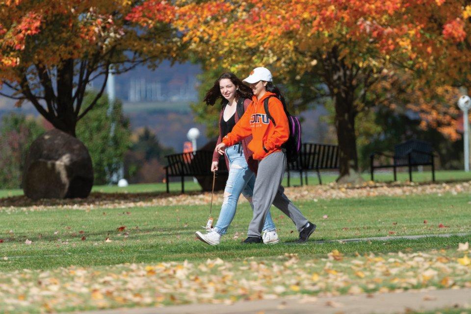 Two students walk across campus amid the red, orange and yellow trees of Autumn.